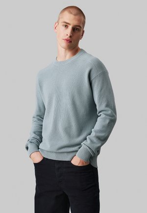 Pull en tricot bleu clair avec un motif texturé, col rond, poignets et ourlet côtelés. Le modèle porte un jean foncé, les mains dans les poches.