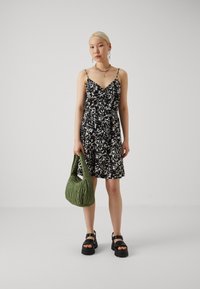Rochie florală alb-negru cu bretele subțiri, talie strânsă și fustă evazată, combinată cu o geantă verde matlasată și sandale negre cu talpă groasă.