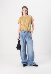 Yellow floral-patterned t-shirt, relaxed-fit blue denim jeans, black handbag, and black shoes. Model poses with hands in pockets.