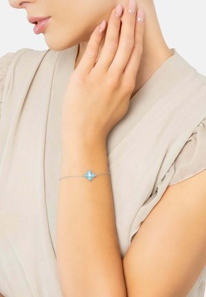 Hopeinen rannekoru, jossa on kukkadesign ja neljä sinistä kiveä. Rannekoru on herkkä ja se lepää vaalealla ihonsävyllä.