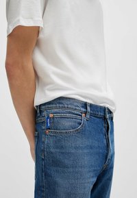 Blue denim jeans with a regular fit, featuring orange stitching, a blue branded tag on the back pocket, and a soft white cotton t-shirt.