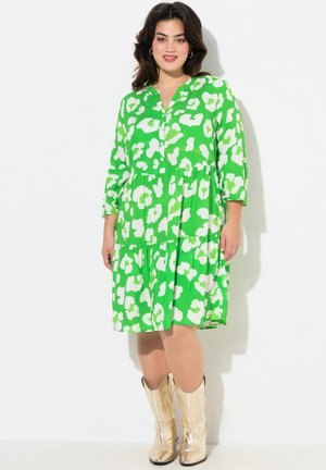 Vestido verde con patrones abstractos blancos, con botones en la parte delantera, mangas de tres cuartos, diseño en capas, combinado con botas de vaquero doradas brillantes.