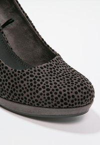 Chaussure en daim gris avec un bout arrondi, présentant un motif texturé à pois noirs. La semelle est plate et conçue en matériau lisse.