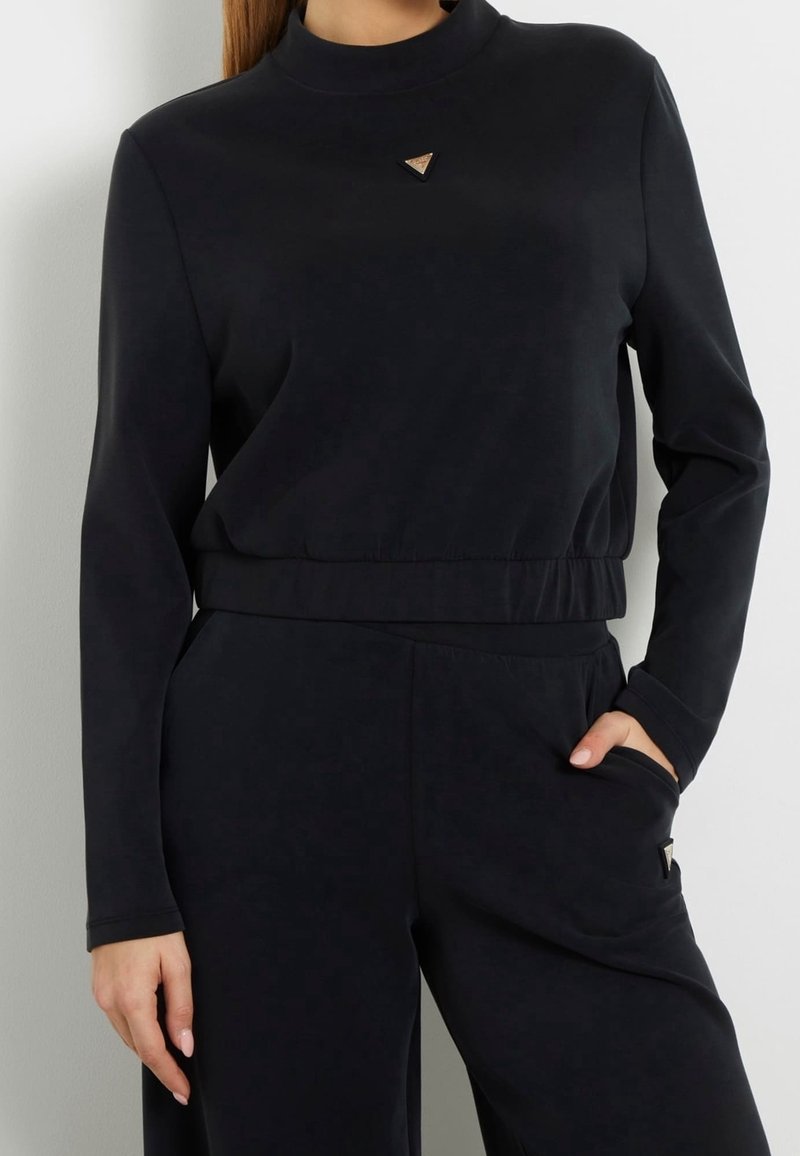 Femme portant un haut noir à manches longues et un pantalon noir assorti, avec une main dans la poche et un petit logo triangulaire sur la poitrine.