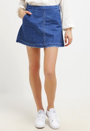 Femme portant une mini-jupe en jean bleu avec un ourlet effiloché, un haut blanc à manches longues et des baskets blanches, debout avec une main dans la poche.