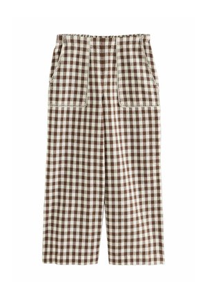 Kobieta ubrana w brązowo-biały kratkowany bezrękawnik i sandały, stojąca na żaglówce z oceanem i czystym niebem w tle.