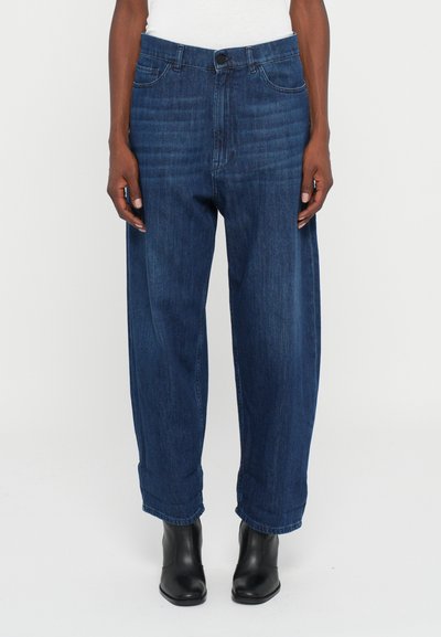 Donkerblauwe losvallende jeans, gedragen met zwarte enkellaarsjes, weergegeven van de taille tot de voeten op een effen witte achtergrond.