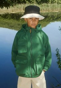 Groene windbreaker jas met twee voren pockets, ritsluiting en een capuchon. Gecombineerd met crèmekleurige broeken en een zwart-wit bucket hat.