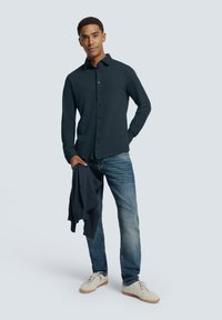 Marineblauw overhemd met lange mouwen en knoopsluiting, gecombineerd met lichtblauwe jeans en beige sneakers. Een donkere trui over de arm gedrapeerd.