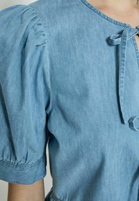 Top in denim azzurro chiaro, caratterizzato da maniche a sbuffo, scollatura rotonda e chiusura frontale con fiocco. Texture morbida con dettagli arricciati sulle maniche.