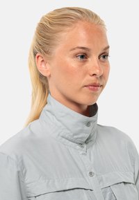 Helles, graues Hemd mit hohem Kragen, das aus einem strukturierten Stoff besteht und zwei Brusttaschen hat. Das Design ist einfach und funktional.