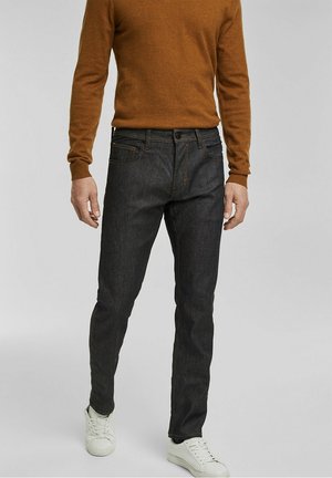 Mężczyzna w dopasowanych ciemnoszarych jeansach, brązowym swetrze z długim rękawem i białych trampkach stoi na jednolitym tle.