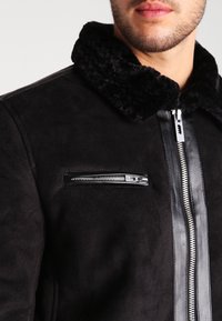 Svart mockajacka med ett pälsfodrat krage, sidofickor och bröstfickor med dragkedja, samt en silverfärgad dragkedjedetalj i mitten. Slät textur och figurnära design.