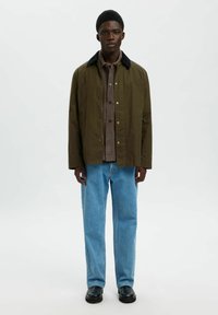 Joven de pie, vistiendo una chaqueta verde oliva con cuello negro, camisa marrón, jeans azul claros y zapatos negros sobre fondo blanco.