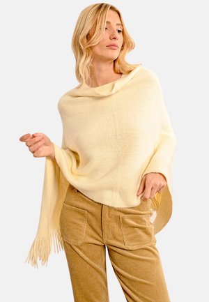 Poncho giallo pallido con un collo alto e vestibilità ampia, caratterizzato da frange lungo l'orlo. Indossato sopra pantaloni di cotone a coste beige.