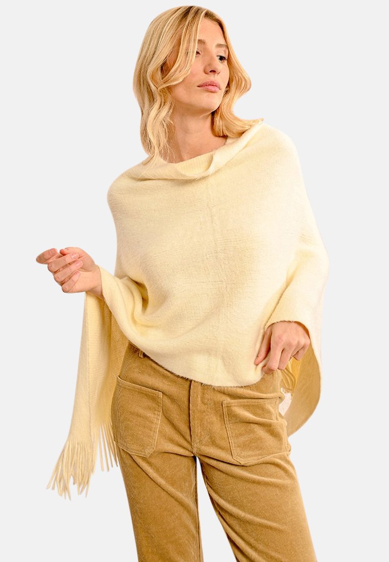 Poncho giallo pallido con un collo alto e vestibilità ampia, caratterizzato da frange lungo l'orlo. Indossato sopra pantaloni di cotone a coste beige.
