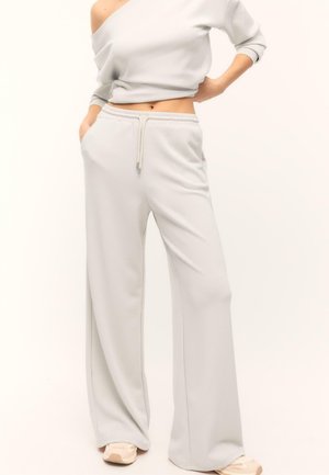 Kobieta w jasno szarym, odsłaniającym ramiona, krótkim topie i szerokich dresowych spodniach z troczkiem, stoi z jedną ręką w kieszeni, nosząc tenisówki.