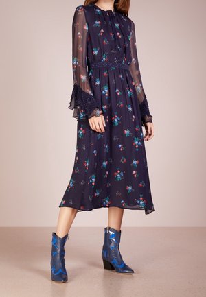 Marineblaues Blumenkleid mit transparenten Ärmeln und tailliertem Schnitt; verfügt über Rüschensäume und mehrfarbige Blumenmuster. Kombiniert mit blauen Absatzstiefeln.