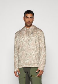 Béžová mikina s kapucňou a camouflage vzorom. Obsahuje predné vrecko a čierne logo hory na hrudníku. Mäkko textúra.