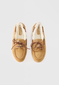 Tan suede moccasin shoes with a fluffy white lining, brown leather laces, and stitched detailing along the edges.