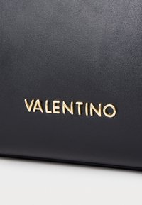 Juodas odinis krepšys su tekstūruota paviršiumi ir aukso „VALENTINO“ logotipo akcentu. Paprastas, elegantiškas dizainas su rafinuotais detalėmis.