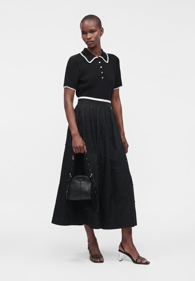 Femme portant un haut noir à manches courtes avec bordure blanche, une jupe midi noire avec de petites décorations, des talons noirs, et tenant un sac à main noir.