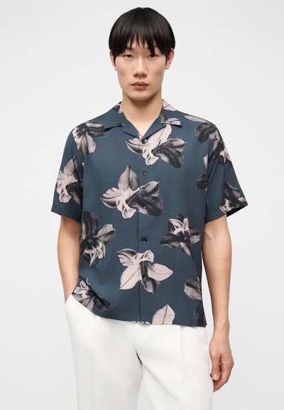Jeune homme portant une chemise bleu foncé à manches courtes avec de grandes impressions florales grises et un pantalon blanc, debout devant un fond uni.