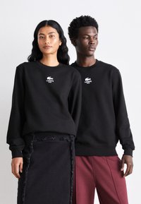 Des sweatshirts noirs à col rond arborent un logo Lacoste brodé en blanc. Ils sont portés avec une jupe noire à franges et un pantalon bordeaux. Fond neutre.