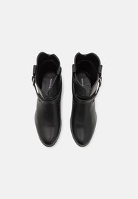 Bottines en cuir noir avec un bout pointu, des bords festonnés et une sangle à boucle réglable. Texture lisse avec des détails de design minimal.