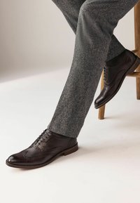 Chaussures brogue en cuir marron foncé à bout cap, avec des détails perforés et des lacets, assorties à un pantalon texturé gris.