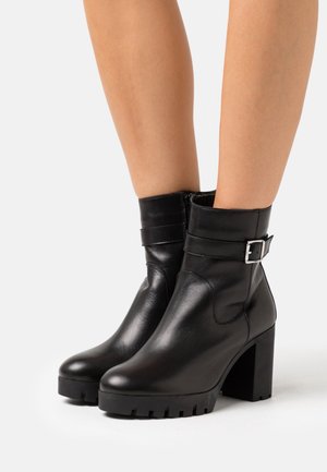 Botas de tobillo de cuero negro con tacón ancho, que cuentan con una correa ajustable con una hebilla de plata y suela de goma texturizada.