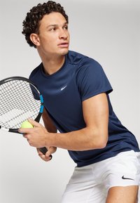Camiseta atlética azul-marinho de mangas curtas, com o logo da Nike no peito, combinada com calções brancos, segurando uma raquete de ténis e uma bola.