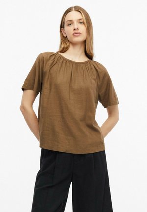 Vrouw met lang blond haar, die een losse bruine korte mouw top en zwarte broek draagt, staat met handen achter haar rug.