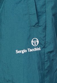 Tyrkysový textil s hladkým povrchom, s bielou vyšívanou značkou a názvom "Sergio Tacchini." Obsahuje detail bočnej vrecka.
