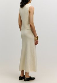 Strick-Zweiteiler in sanftem Creme, bestehend aus einem ärmellosen Oberteil und einem bodenlangen Rock. Glatte Textur mit dezenten Musterdetails. Schwarze Schuhe.