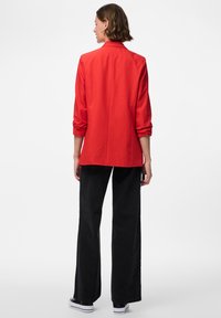 Červený blazer s dlouhými rukávy, klopovým límcem a rovný střih. Spárován s širokými černými kalhotami a bílými teniskami. Hladká textura tkaniny.