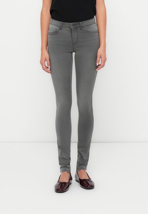 Person trägt schmal geschnittene graue Jeans, schwarzes Oberteil und glänzende dunkelrote Ballerinas, steht vor einem schlichten weißen Hintergrund.