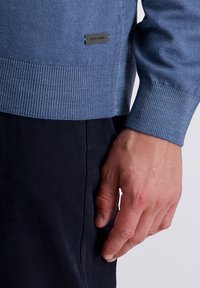 La mano di un uomo accanto a pantaloni blu navy, indossa un maglione azzurro a coste con una piccola etichetta nera Pierre Cardin vicino all'orlo.