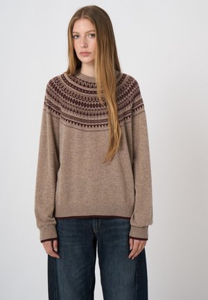 Jonge vrouw met lang blond haar, die een beige gebreide trui met patroon en donkerblauwe spijkerbroek draagt, staand tegen een effen lichte achtergrond.