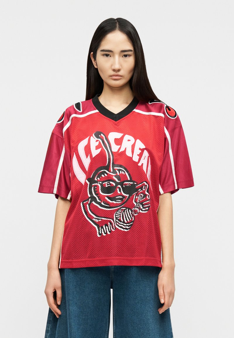 Νεαρή γυναίκα με μακριά σκούρα μαλλιά φοράει μια φαρδιά κόκκινη αθλητική μπλούζα με χαρακτήρα παγωτού που χαμογελά, φοράει γυαλιά ηλίου και μπλε τζιν.