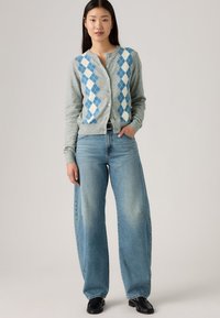 Cardigan gris avec un motif argyle bleu et blanc, doté de boutons sur le devant. Associé à un jean à pattes d'éléphant en denim clair et des chaussures noires.