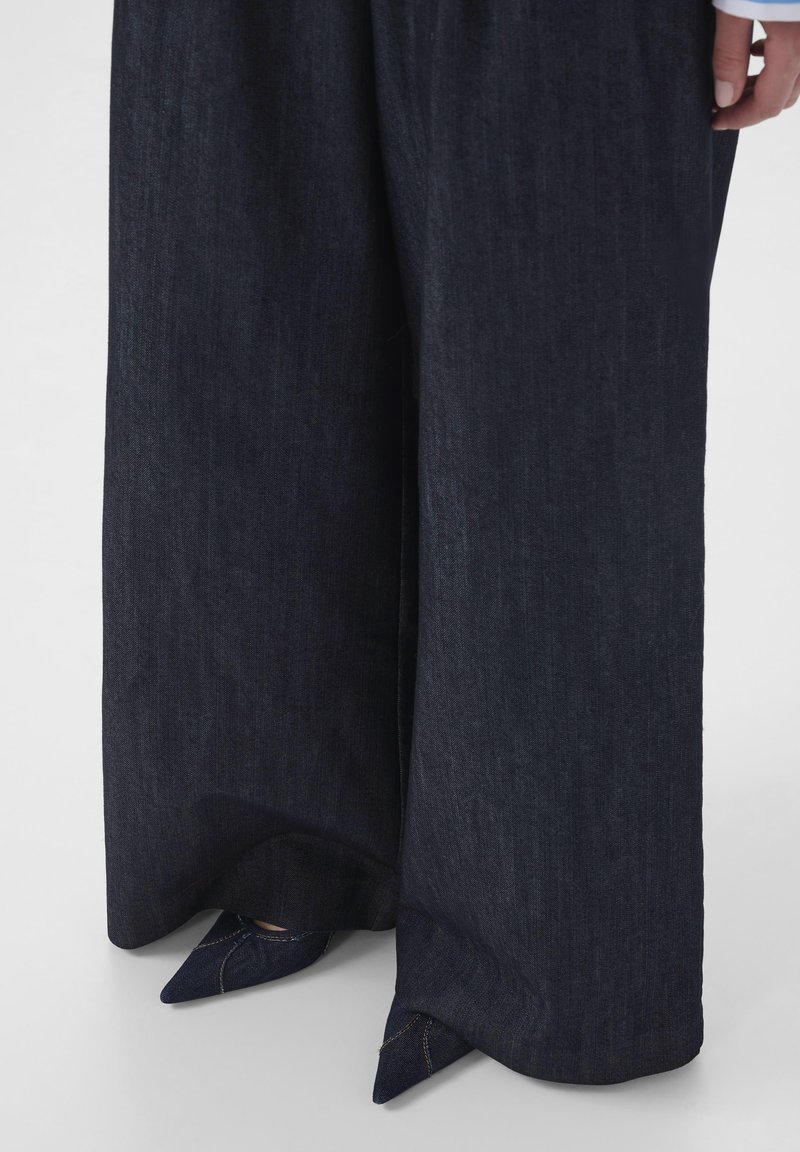 Pantalon large en denim foncé à coupe droite, avec une texture subtile. Associé à des talons pointus bleu marine, présentant un design élégant.