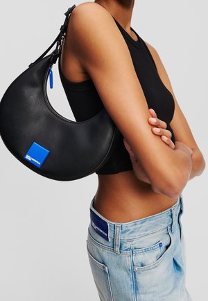 Personne portant un crop top noir et un jean bleu clair avec un sac épaule noir en forme de croissant, orné d'un écusson logo bleu.