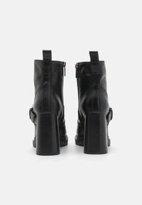 Botas de cuero negras hasta el tobillo con un tacón grueso, cremalleras laterales y un detalle de hebilla. Textura suave con un diseño de rayas verticales en el tacón.