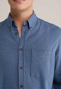 Kék, gombos ing finom halszálka mintával, mellkasi zsebbel és sötét gombdíszítéssel; az anyag textúrája simának és puhanak tűnik.