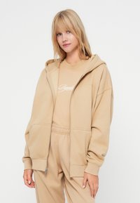 Beige zipsová mikina s voľným strihom, prednými vreckami a rebrovanými manžetami. Nosí sa cez zodpovedajúce béžové tričko s bielym logom.
