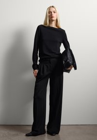 Zwarte trui met lange mouwen, losse zwarte broek en een getextureerde zwarte tas, gerangschikt tegen een neutrale achtergrond. Platte schoenen worden gedragen.