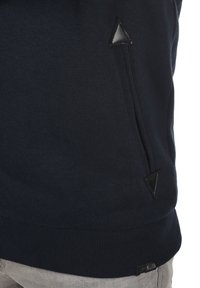 Sweatshirt bleu marine avec un ourlet côtelé, présentant des accents triangulaires en cuir noir près d'une poche zippée et une texture de tissu lisse.