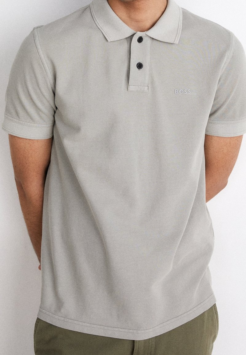 Polo gris clair avec un col classique, deux boutons noirs et un logo 'BOSS' discret. Tissu en coton, manches courtes et ourlet droit.