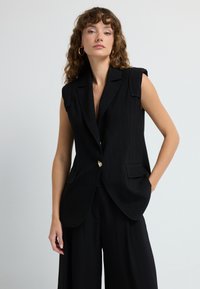 Černá bezrukávová vesta s vykrojeným límcem a jedním zlatým knoflíkem. Má dvě přední kapsy a hladkou, strukturovanou tkaninu.